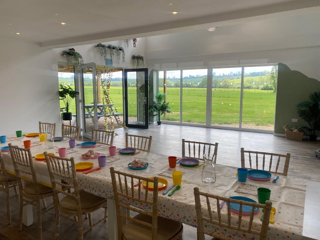 Long table laid for guests in a bright barn, with colourful place settings and views of green fields through full-height windows.